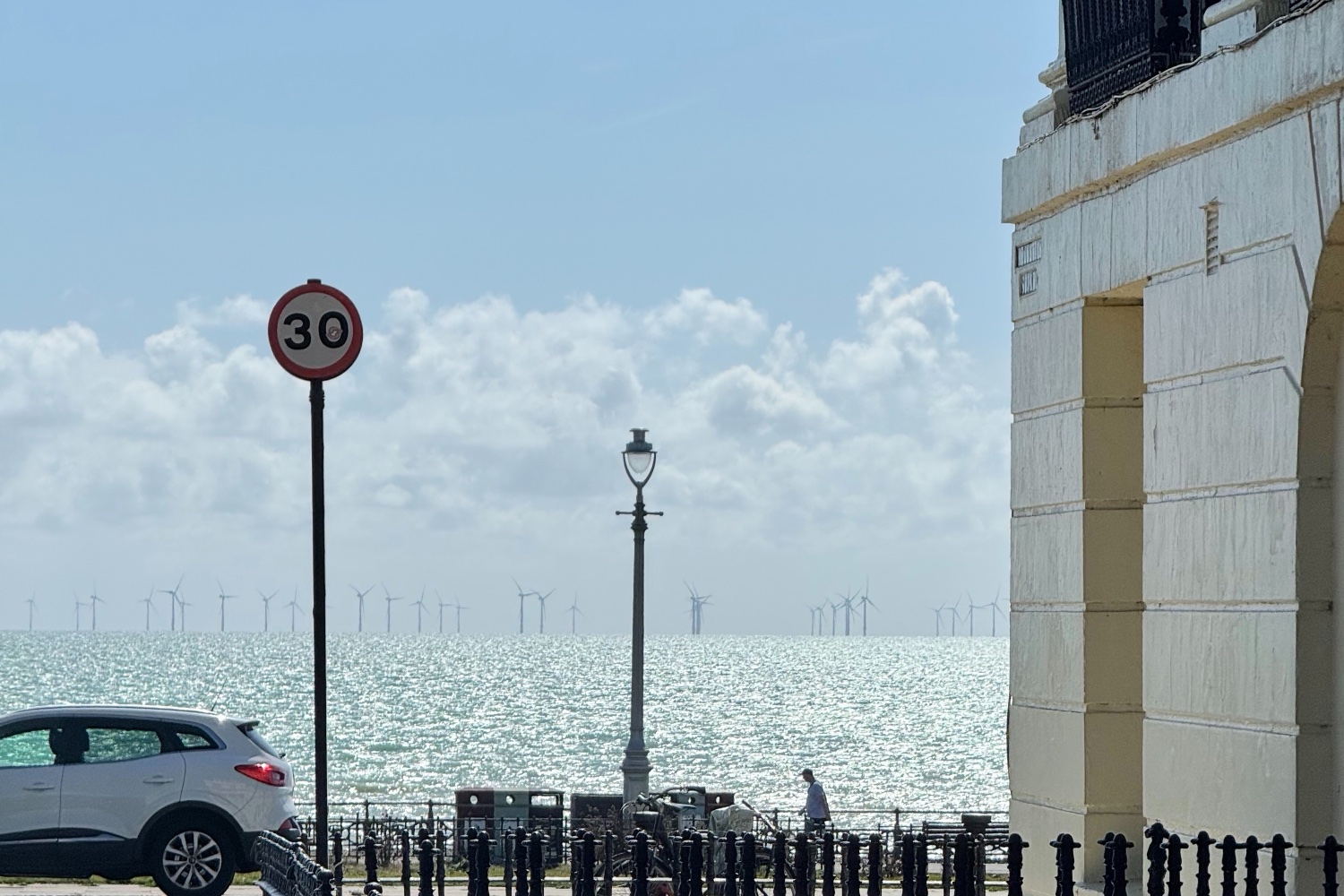 Brighton seafront viewed past black railings and a Regency building, with a lamp post and offshore wind turbines across the sea